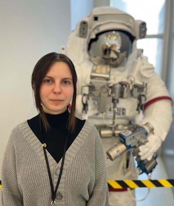 Eva T. from Ohio Connections Academy in a black turtleneck shirt and brown sweater standing in front of a space suit display at NASA Glenn Research in Cleveland. 